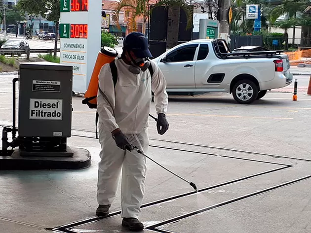 Profissional realiza desinfecção em área de posto de gasolina, usando equipamento de proteção e pulverizador.